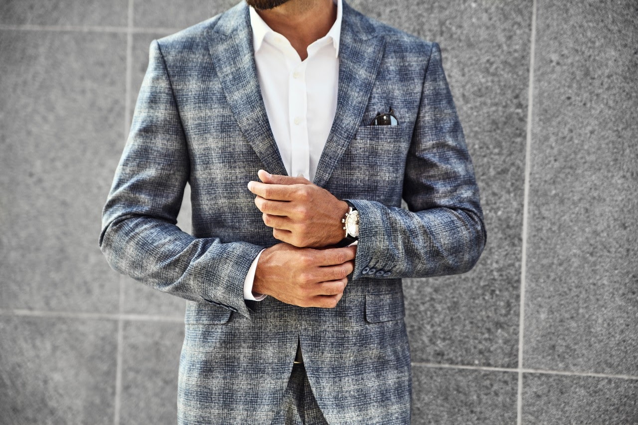 A man wearing a gray plaid suit adjusts his timepiece while standing against a gray tile wall