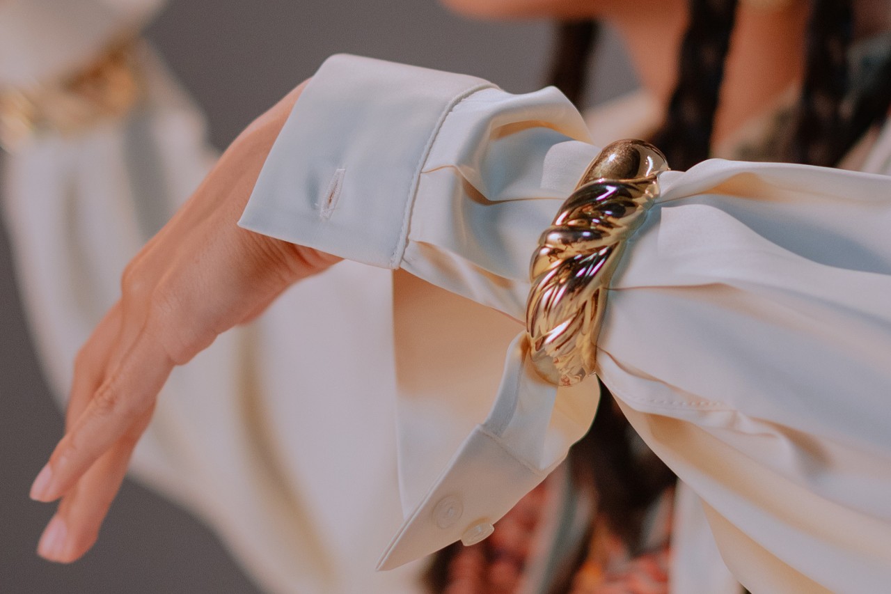 a person’s extended arm clad in a white blouse sleeve and chunky gold bangle bracelet