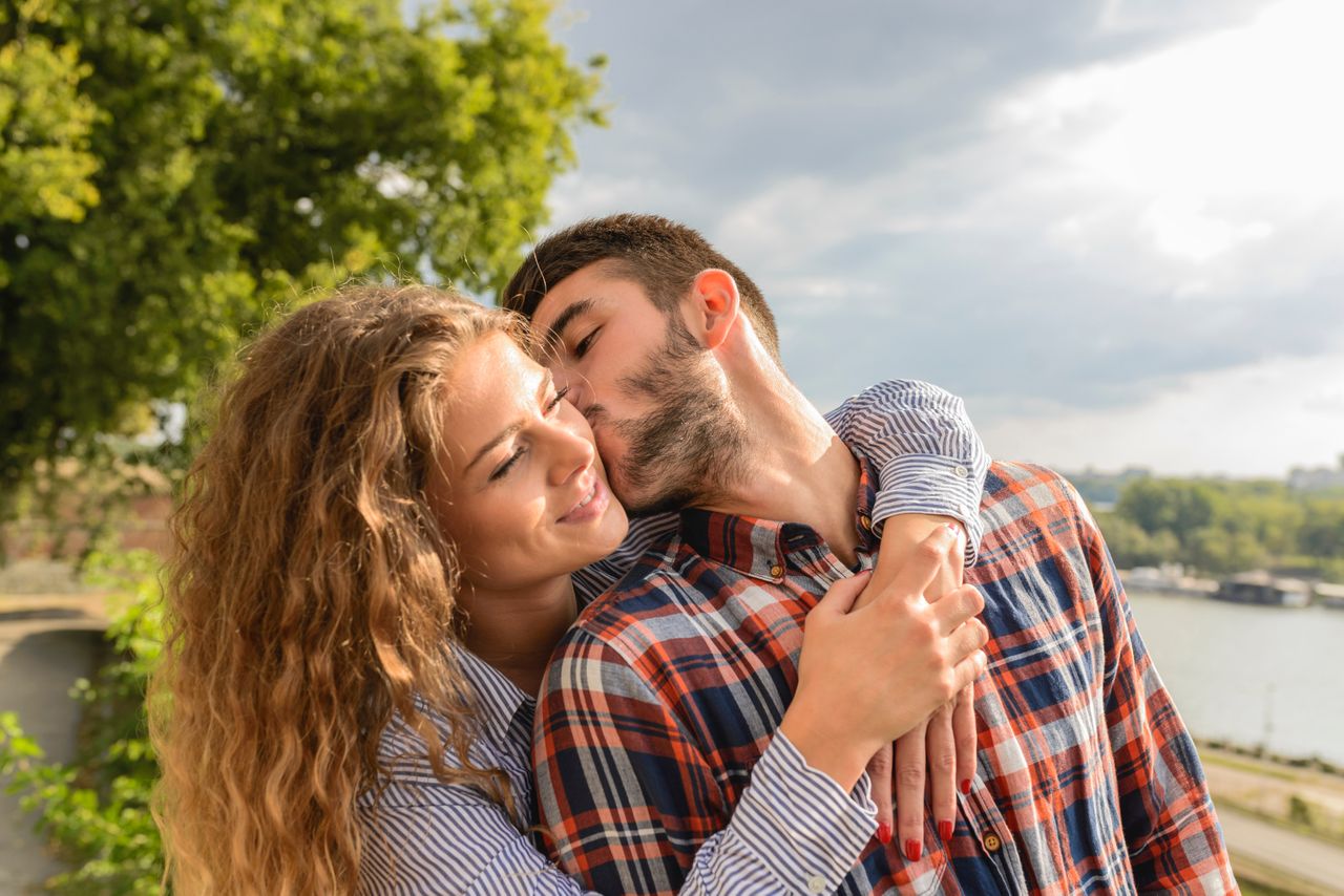 A couple by open water, woman embracing man from behind as he kisses her cheek.