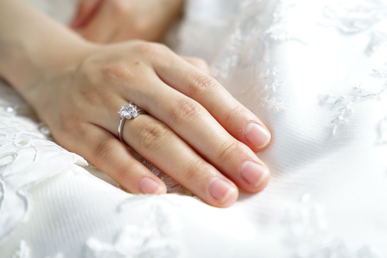 Close up of a bride&rsquo;s hand showcasing the solitaire engagement ring resting oa white lace gown.