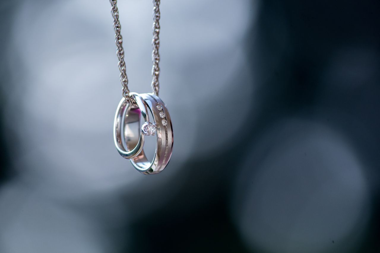 An engagement ring and a wedding band hanging on a silver chain with a blurred background.