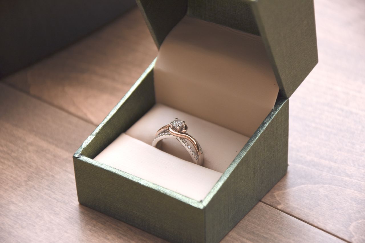 A mixed-metal engagement ring inside a green ring box on a wooden table.