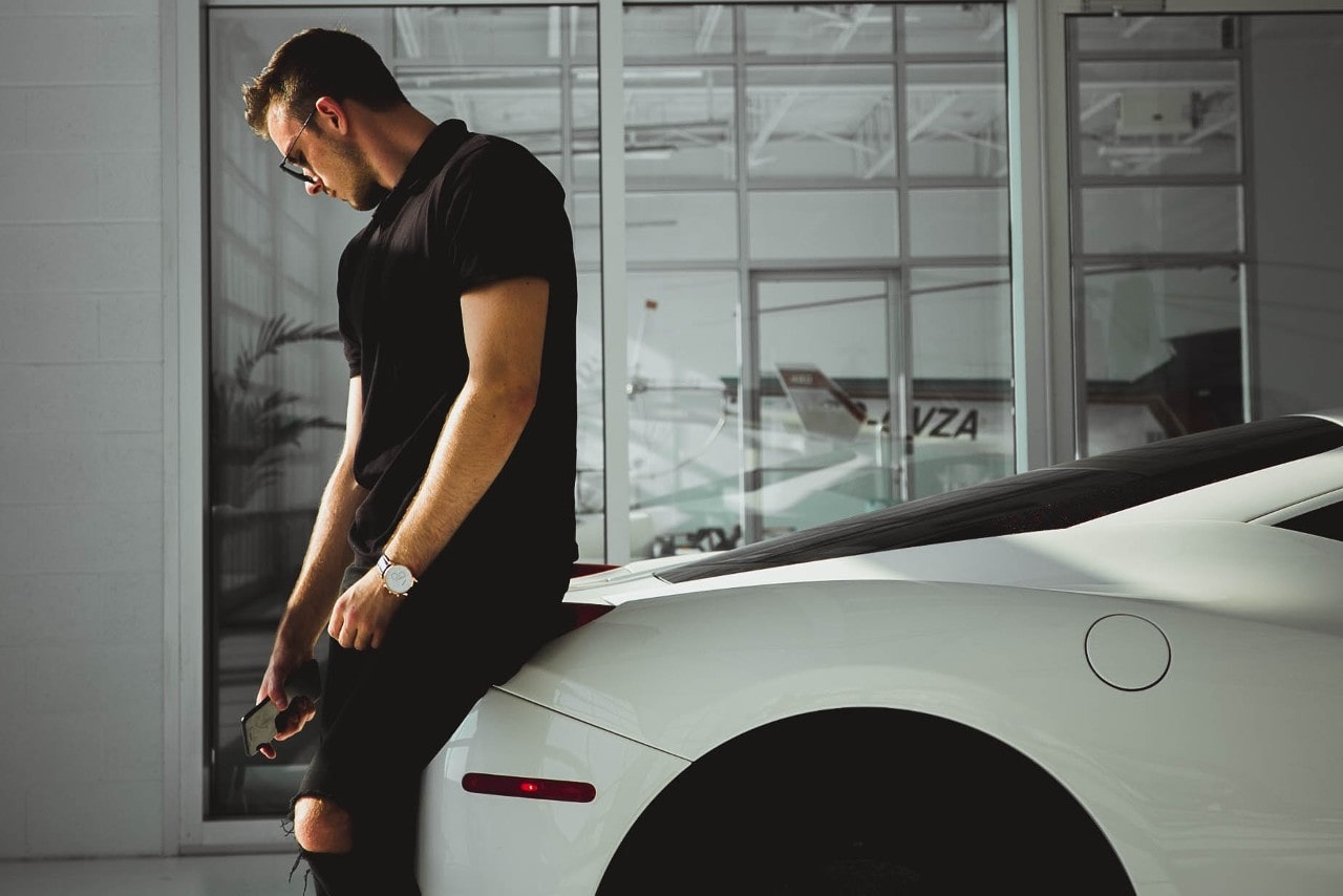 A man leans on a sports car, showing off his casual timepiece.