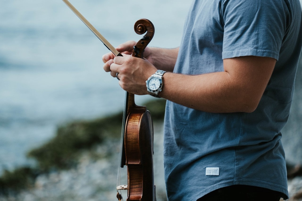 A casual man in a blue shirt holds a violin on the shore of a river, wearing a timepiece.