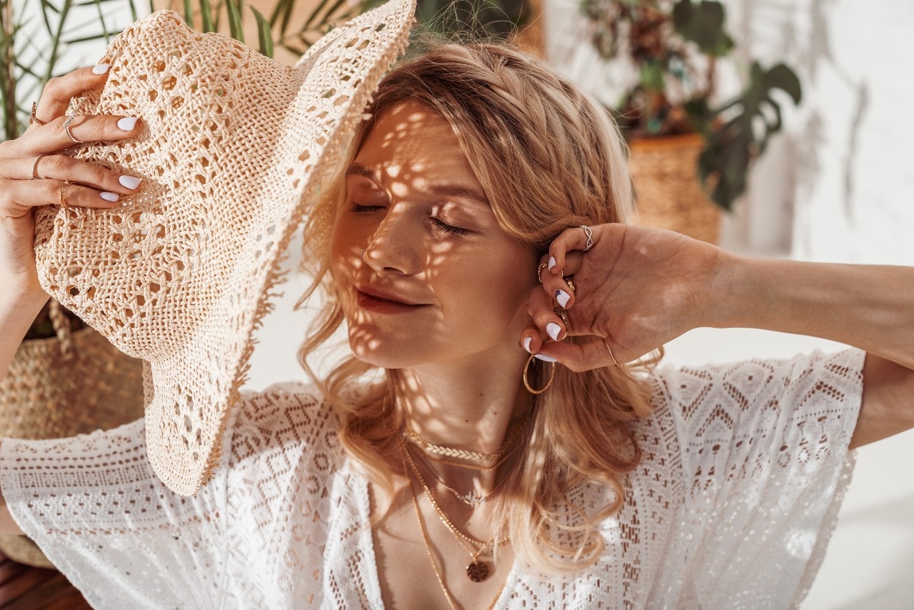 A woman in a white embroidery blouse and hat smiles against the sunlight, showcasing layered yellow gold necklaces, rings, and hoop earrings.