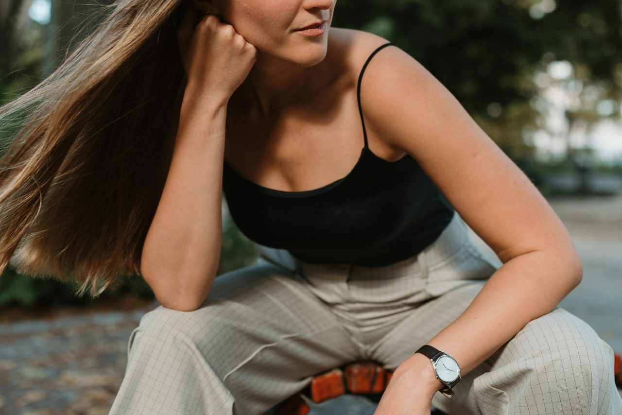 A close-up of a casually dressed young woman sitting on a red outdoor bench, a minimalist luxury watch on her left wrist.
