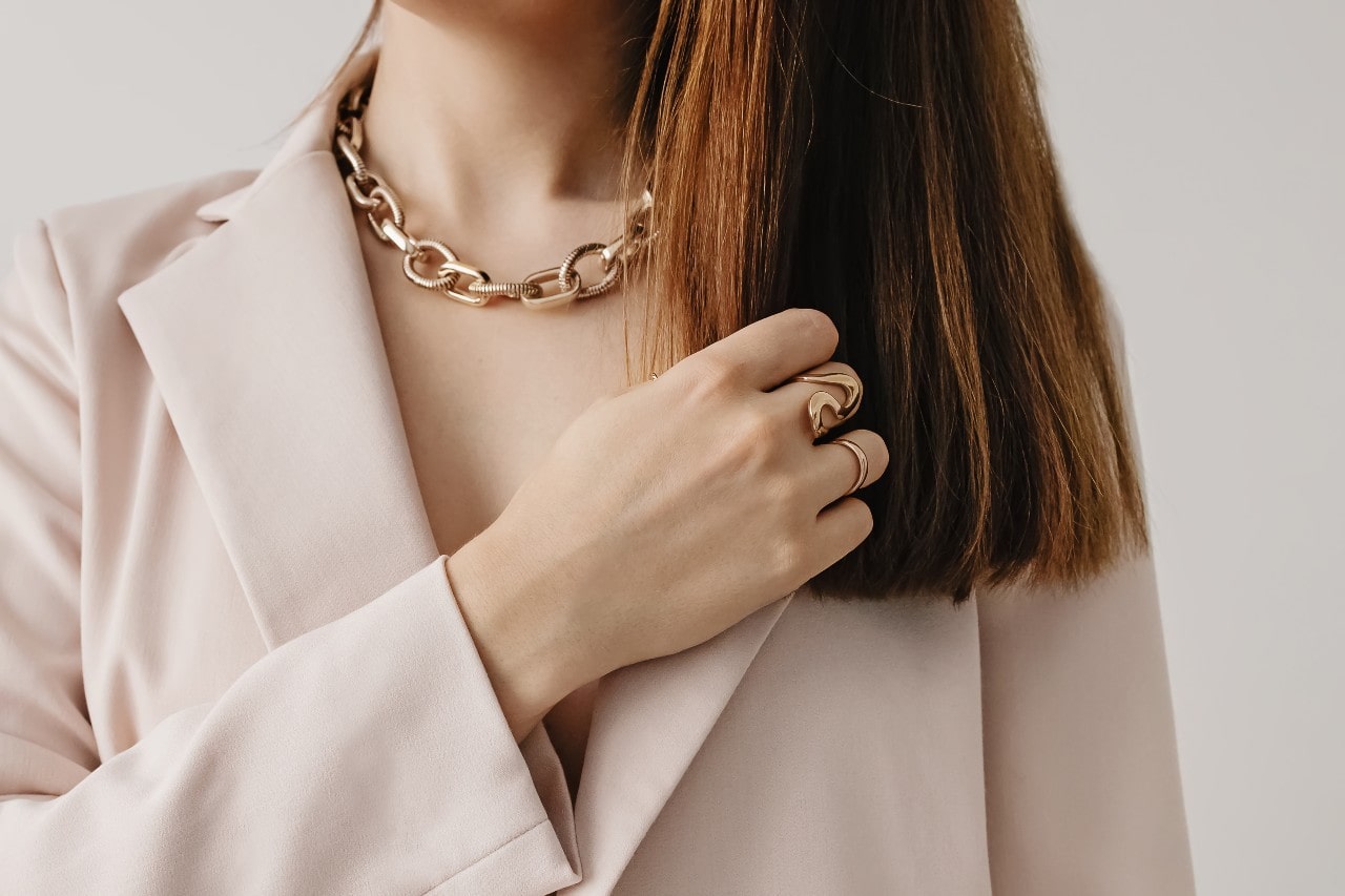 A woman in a beige blazer showcases a yellow gold chain necklace and fashion rings.