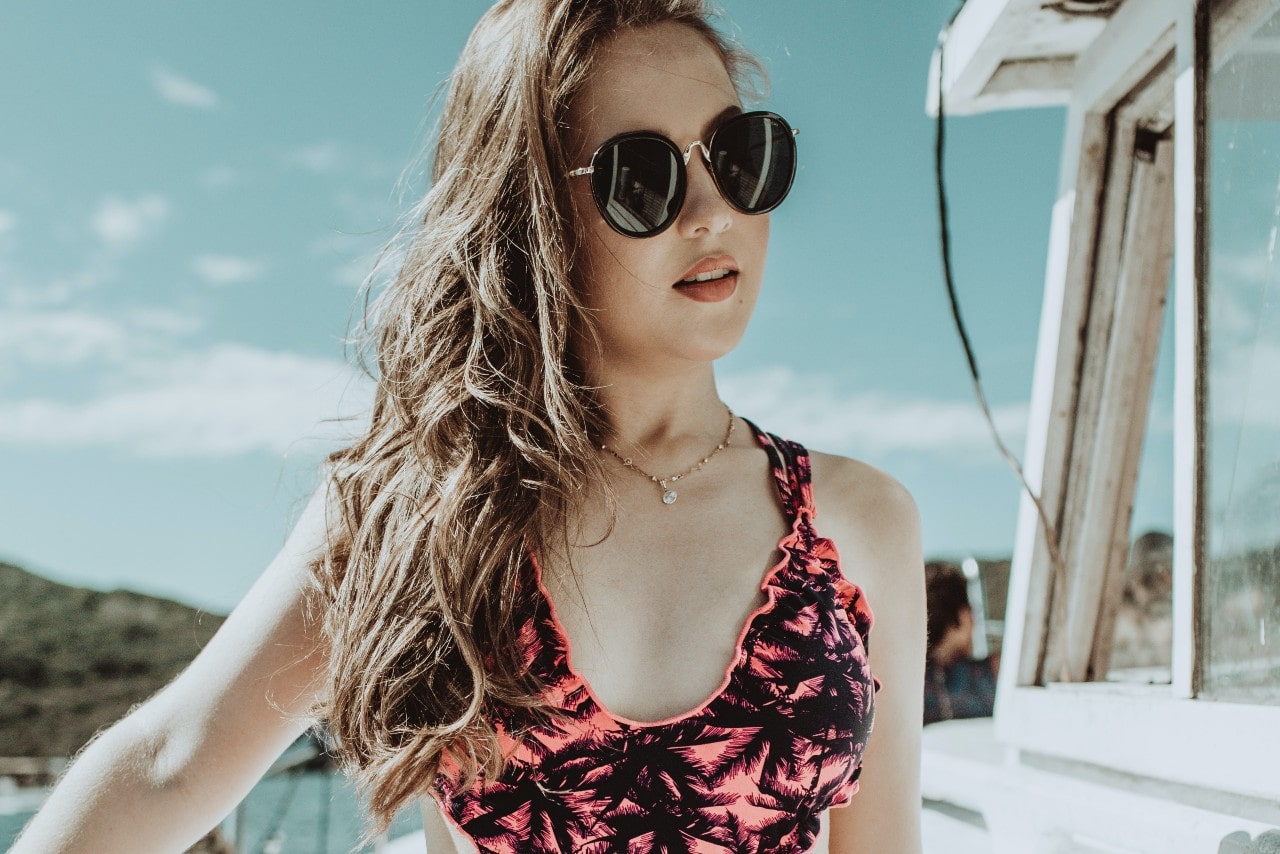 A woman in a printed pink swim bra and sunglasses gazes away, showcasing a yellow gold pearl necklace.