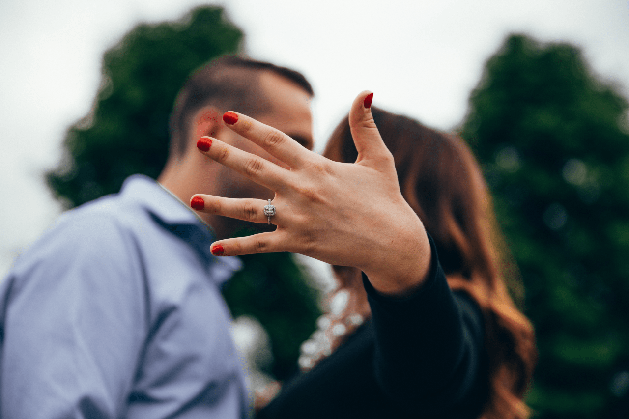 A man and woman kissing, their kiss being blocked by the woman holding up her hand, showcasing her gorgeous engagement ring.