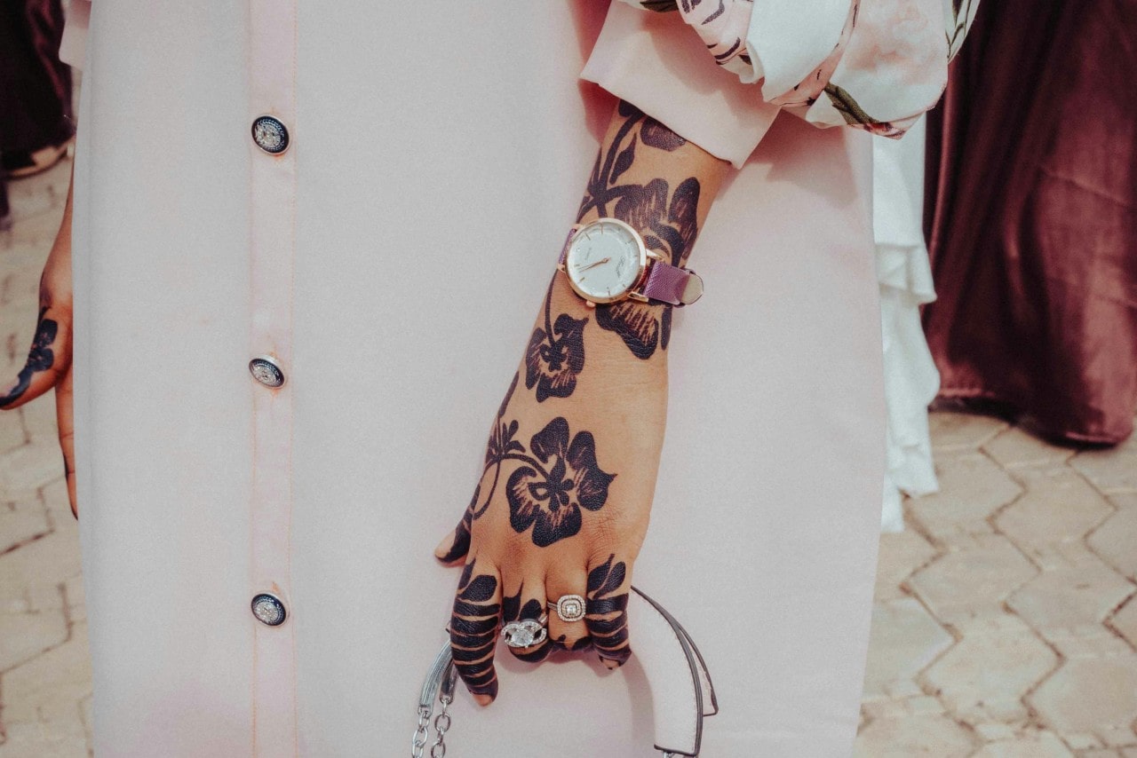 A woman wearing floral henna designs on her hand showcases a gold wristwatch with rose stripe, statement rings, and a small white handbag against a pale pink buttoned dress.