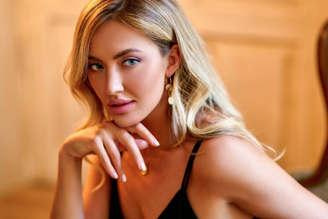 A blonde woman in a black top wearing drop earrings rests her chin on her hand against a softly blurred background.