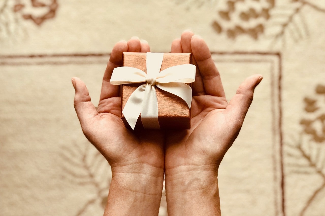A top-down view of a person holding a small jewelry gift box wrapped in a cream-colored ribbon in their hands.