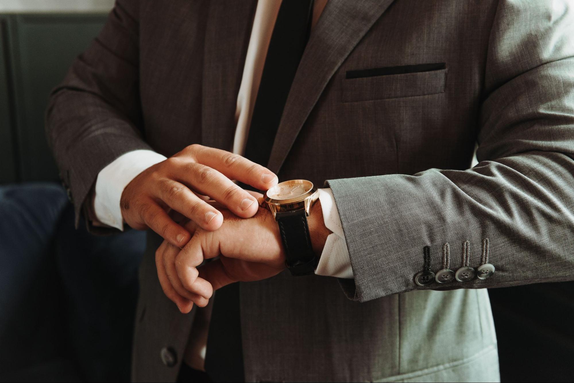 A close-up of a man in a grey suit looking at his black leather dress watch with a gold-plated bezel.
