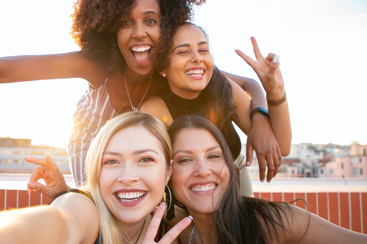 Four smiling women posing for a photograph at the beachside, two showing peace signs.