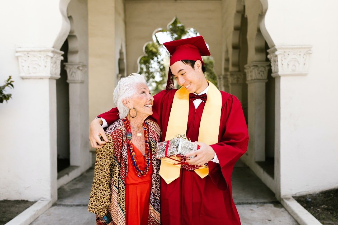 A graduate in a red cap and gown with his grandmother, holding a gift in his hands.