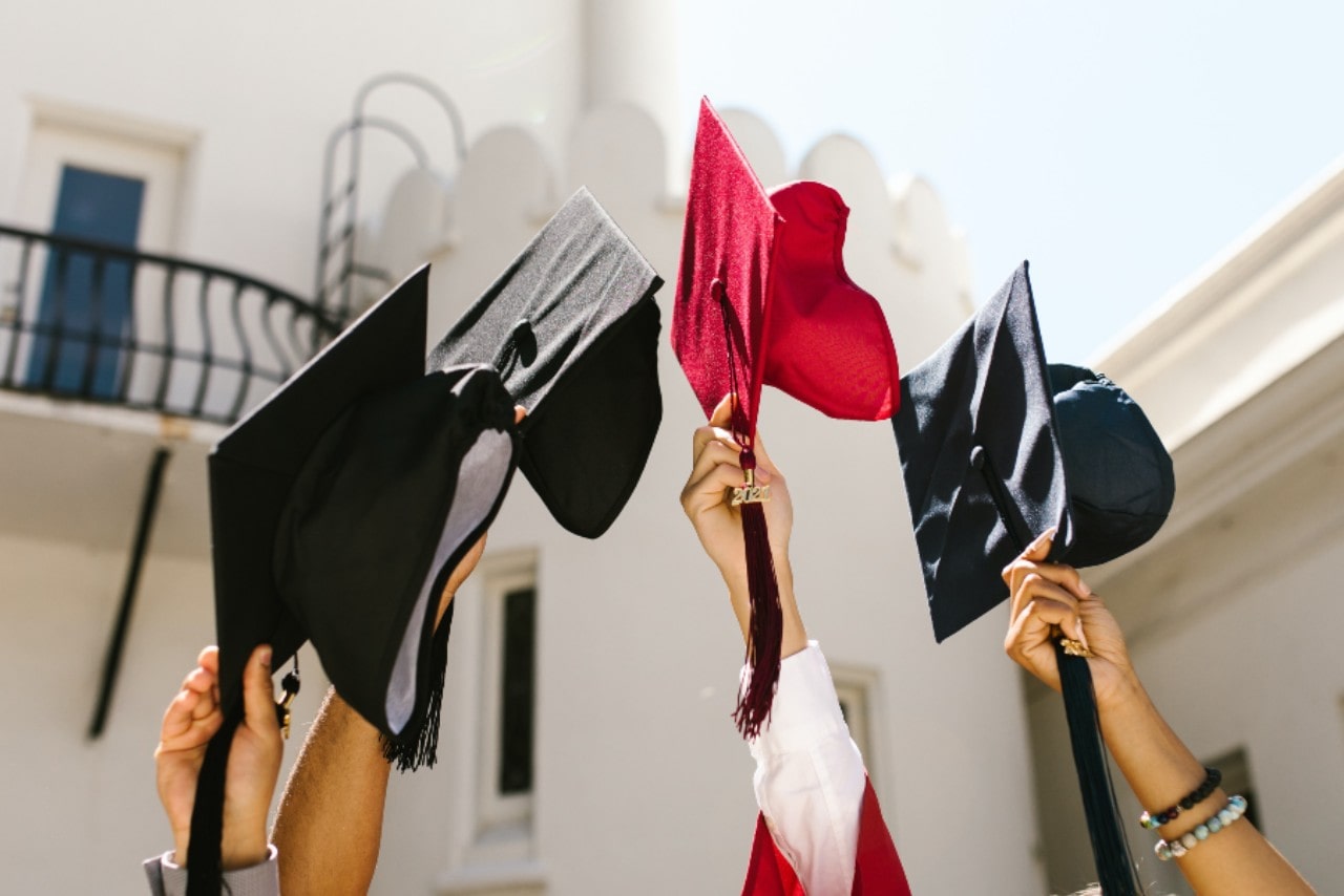 Several hands holding up black and red graduation caps in front of a white building.
