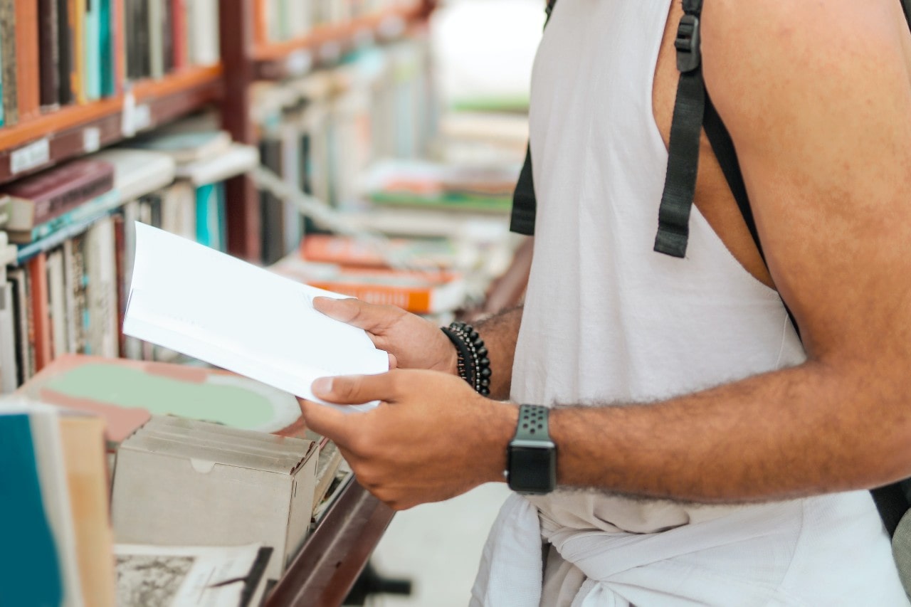 A person in a sleeveless shirt with bracelets, a luxury watch, and a backpack browses books in a library.