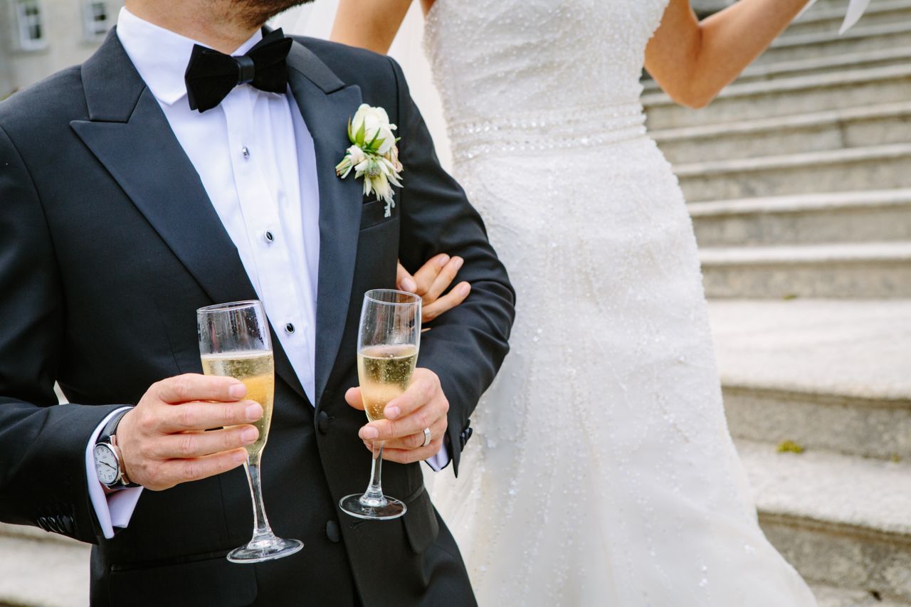 The bride in a beaded white gown links arms with a groom in a black tuxedo wearing a luxury watch and holding two champagne glasses, standing on steps in a joyful celebration.