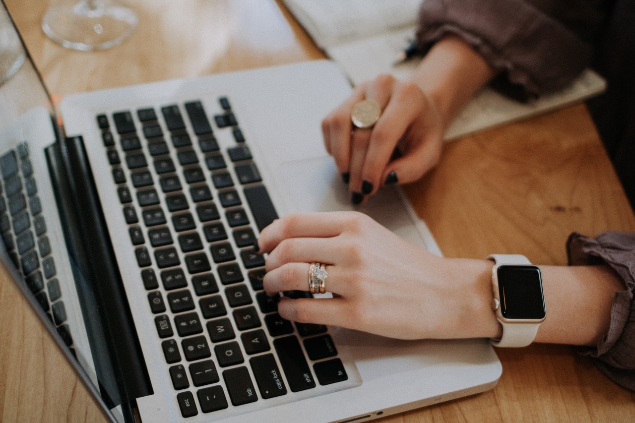 A person with rings and a smartwatch types on a laptop resting on a wooden table.