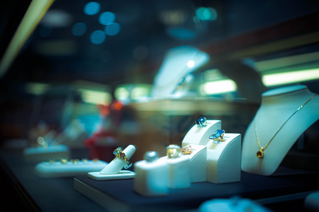 A jewelry store display case showcasing bold gemstone rings in gold settings and a delicate gold pendant necklace, all arranged under soft lighting.