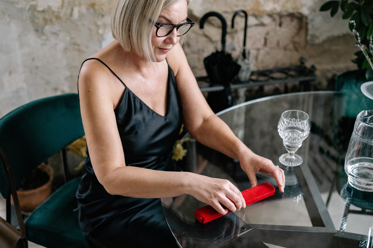 A woman with glasses and a black dress sits at a glass table, unrolling a red velvet rectangular box.
