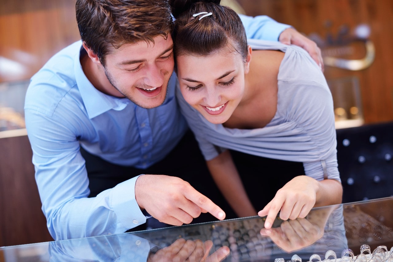 Smiling couple in a jewelry store, leaning over a glass counter, eagerly pointing at rings.