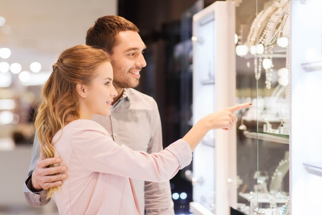 A happy couple glancing at the jewelry store window, with a woman pointing out and a man hugging her with one hand.