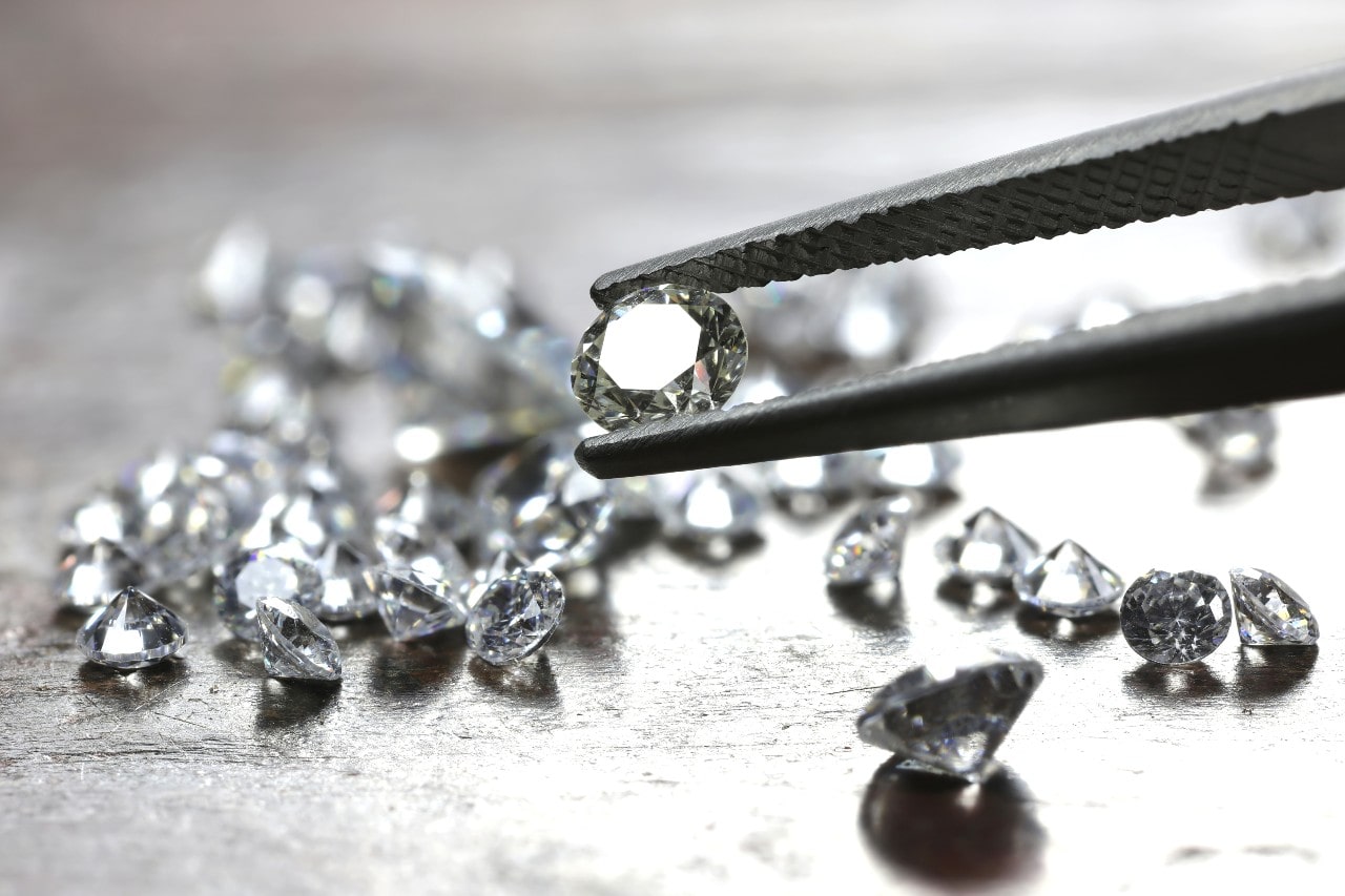 A macro shot of tweezers holding a loose round diamond above a scattered collection of sparkling diamonds.