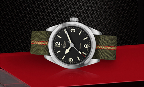 A close-up of a TUDOR Ranger watch with a green fabric strap displayed on its side on a red and black background.