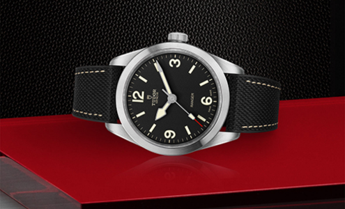 A close-up of a TUDOR Ranger watch with a black fabric strap displayed on its side on a red and black background.