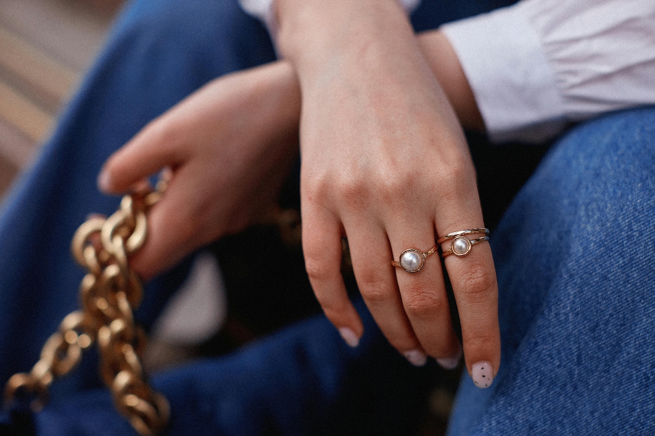 A woman sitting holding the gold chain of her purse in one hand with a focus on her yellow gold and pearl rings on her other hand