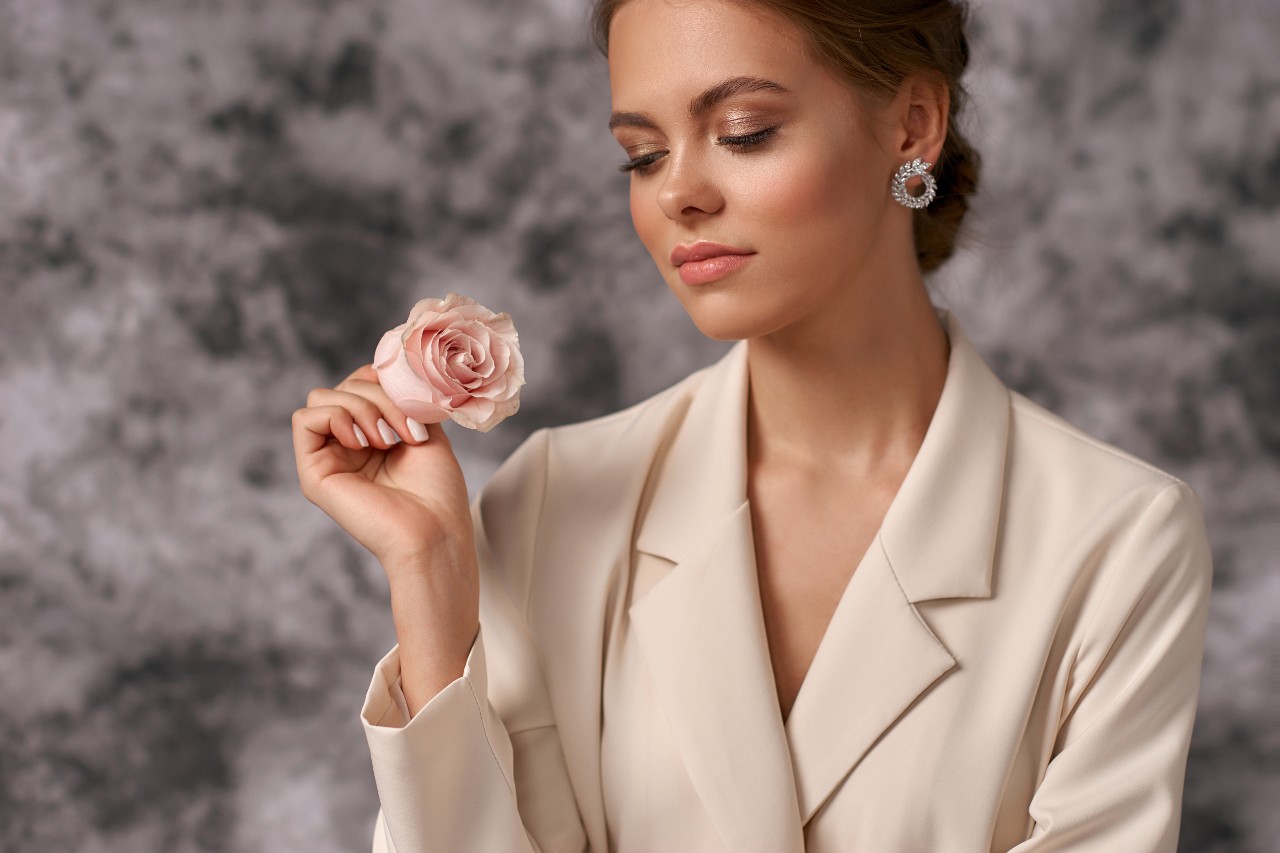 A woman in a cream blazer and sparkling diamond earrings holding a pink rose.