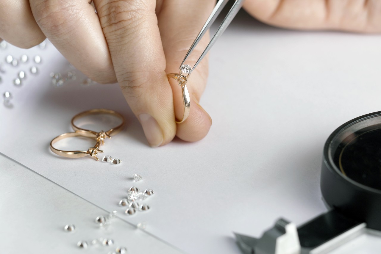 A close-up of a jeweler’s hand using tweezers to place a diamond on a gold ring, surrounded by loose diamonds and two completed rings.