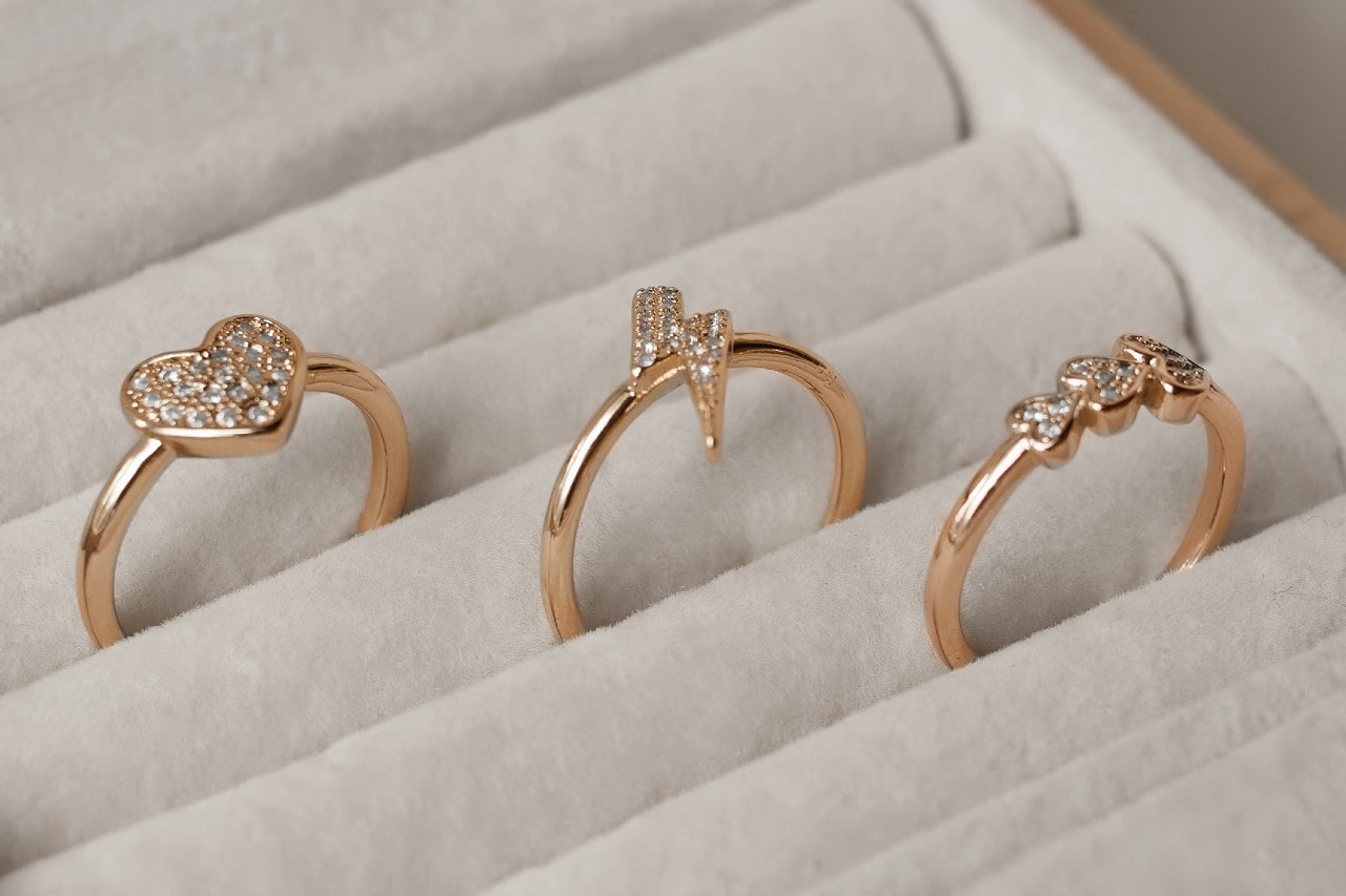 A close-up of 3 yellow gold fashion rings with sparkling diamonds in a different shape – a heart, a lightning bolt, and a trio of round stones – rest in individual slots within a light-colored jewelry display.