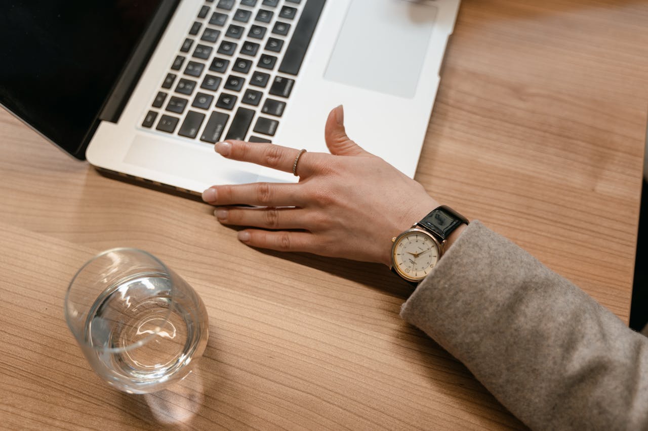 A top-down view of a person’s hand, adorned with a luxury watch and subtle fashion ring, as they type on a laptop on a wooden desk.
