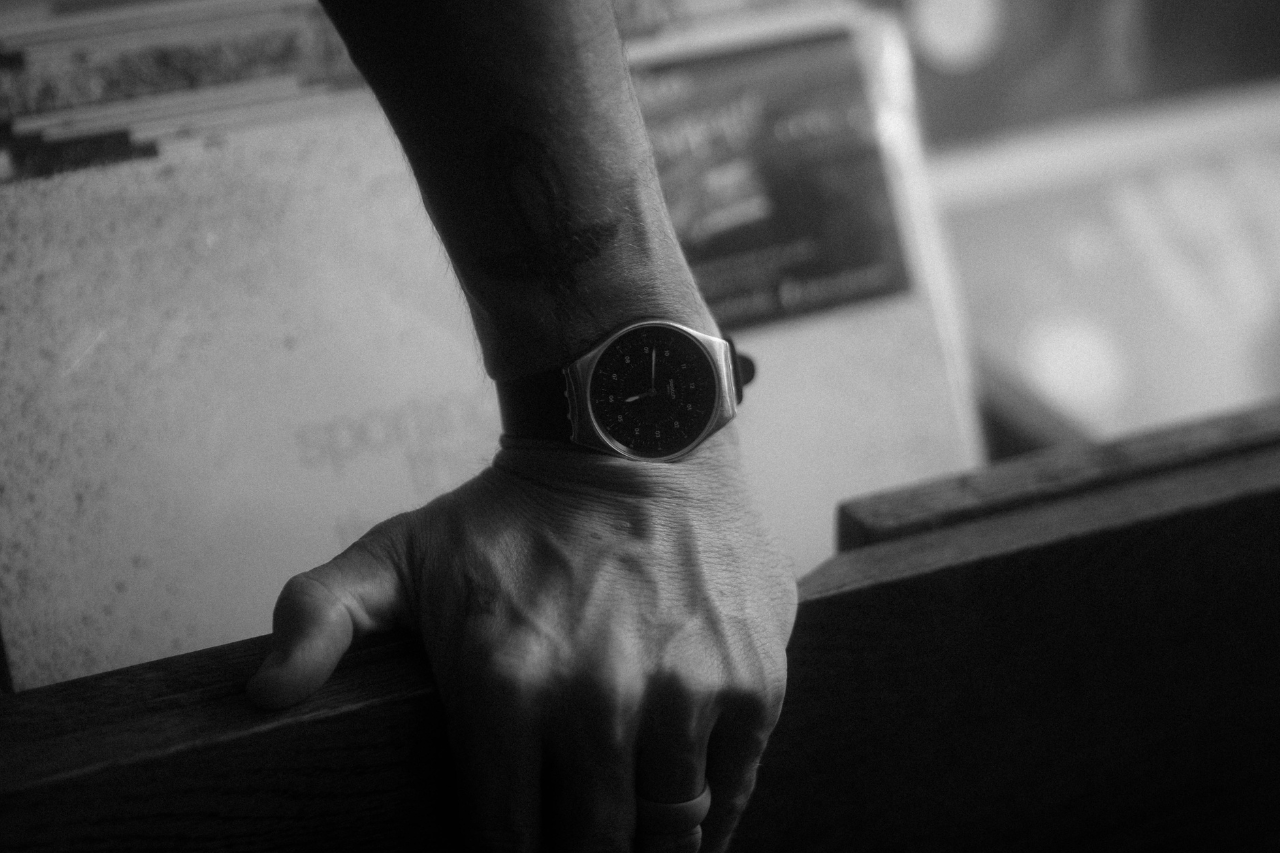 A close-up of a man’s hand, a luxury watch on his wrist, resting atop a wooden vinyl display shelf.