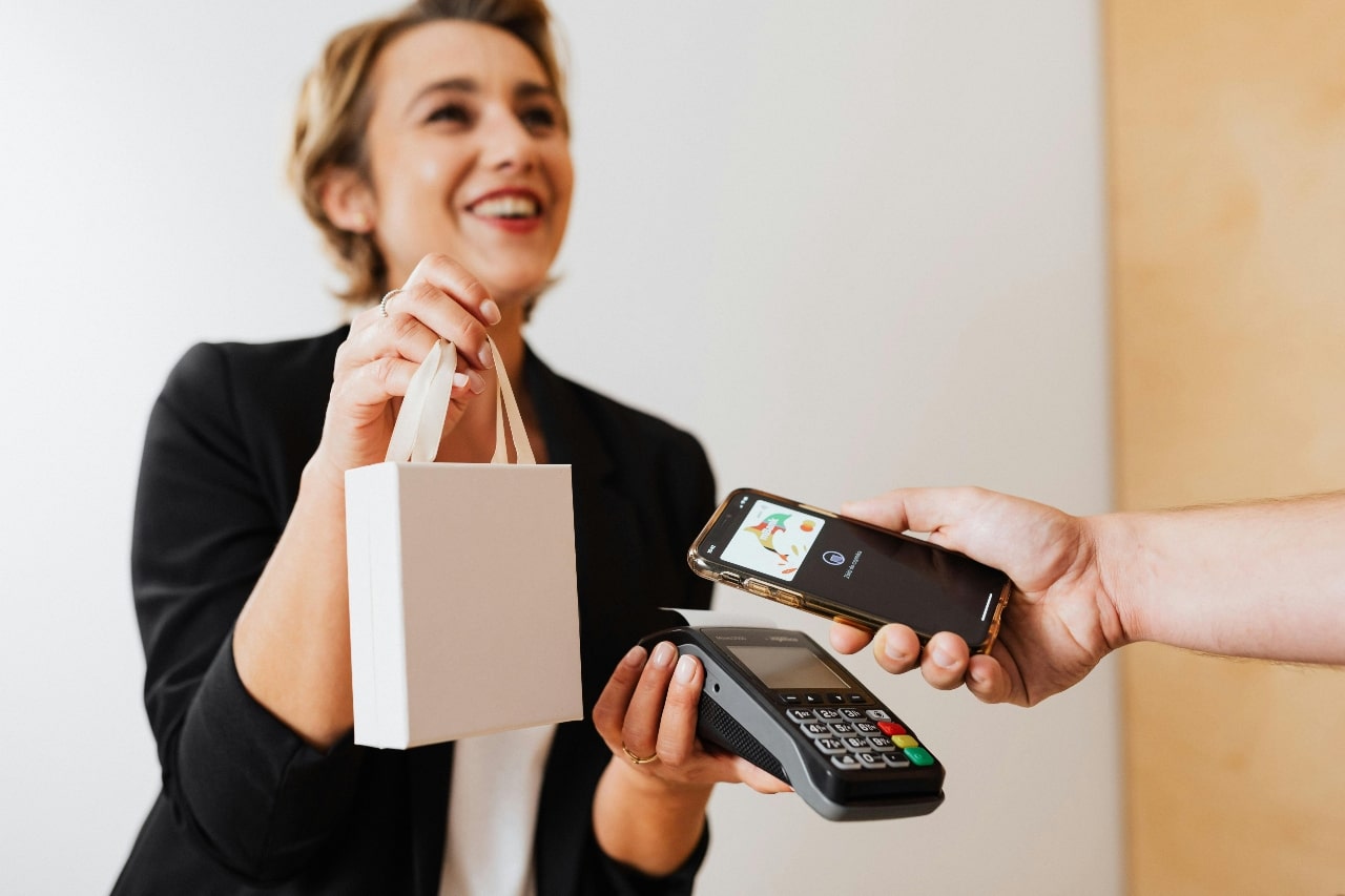 A smiling jeweler hands a small gift bag to their customer while they use a smartphone for contactless payment.
