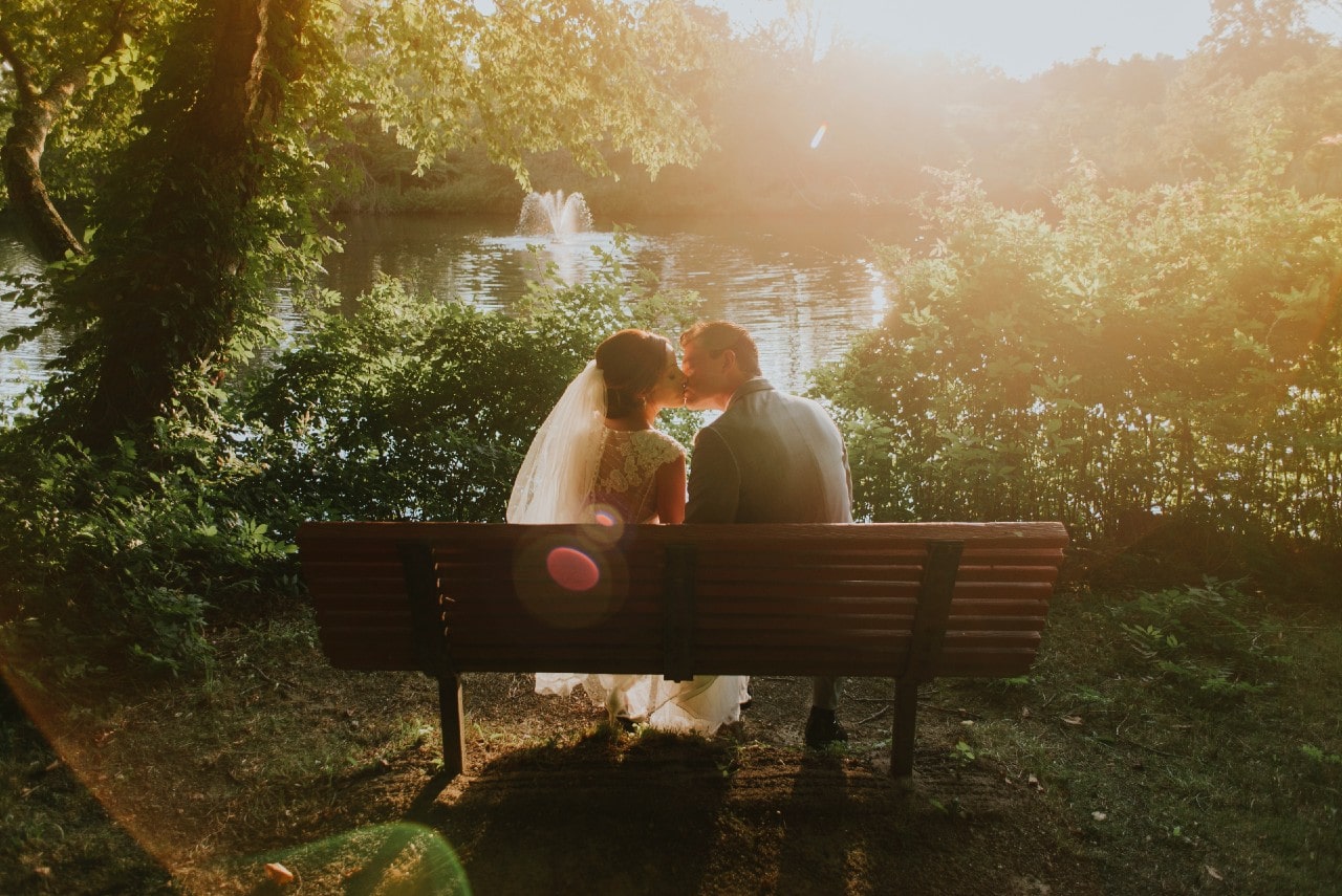 A couple in wedding attire kissing on a bench with a soft and romantic filter.