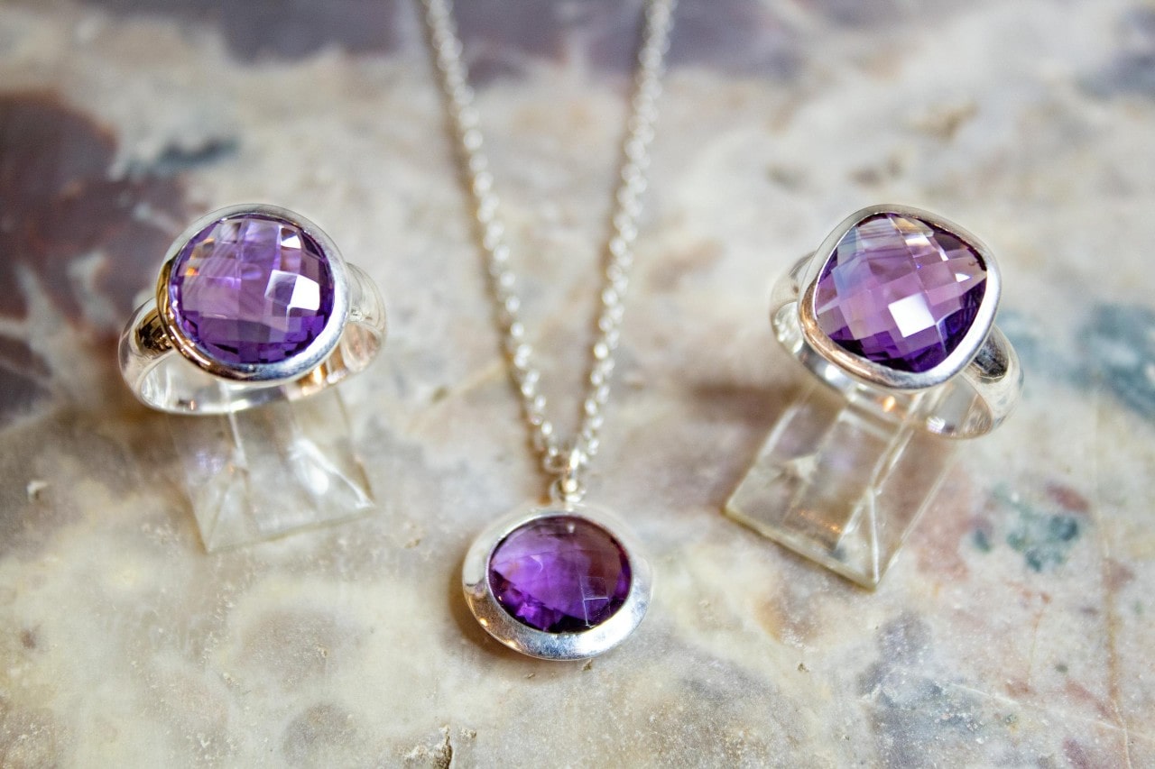 Elegant jewelry set with faceted purple amethyst stones displayed on a textured beige surface.