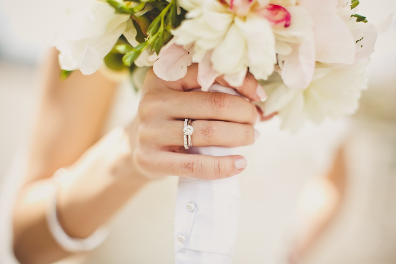 Close-up of a hand in a white dress with a solitaire diamond engagement ring holding a bouquet of flowers.