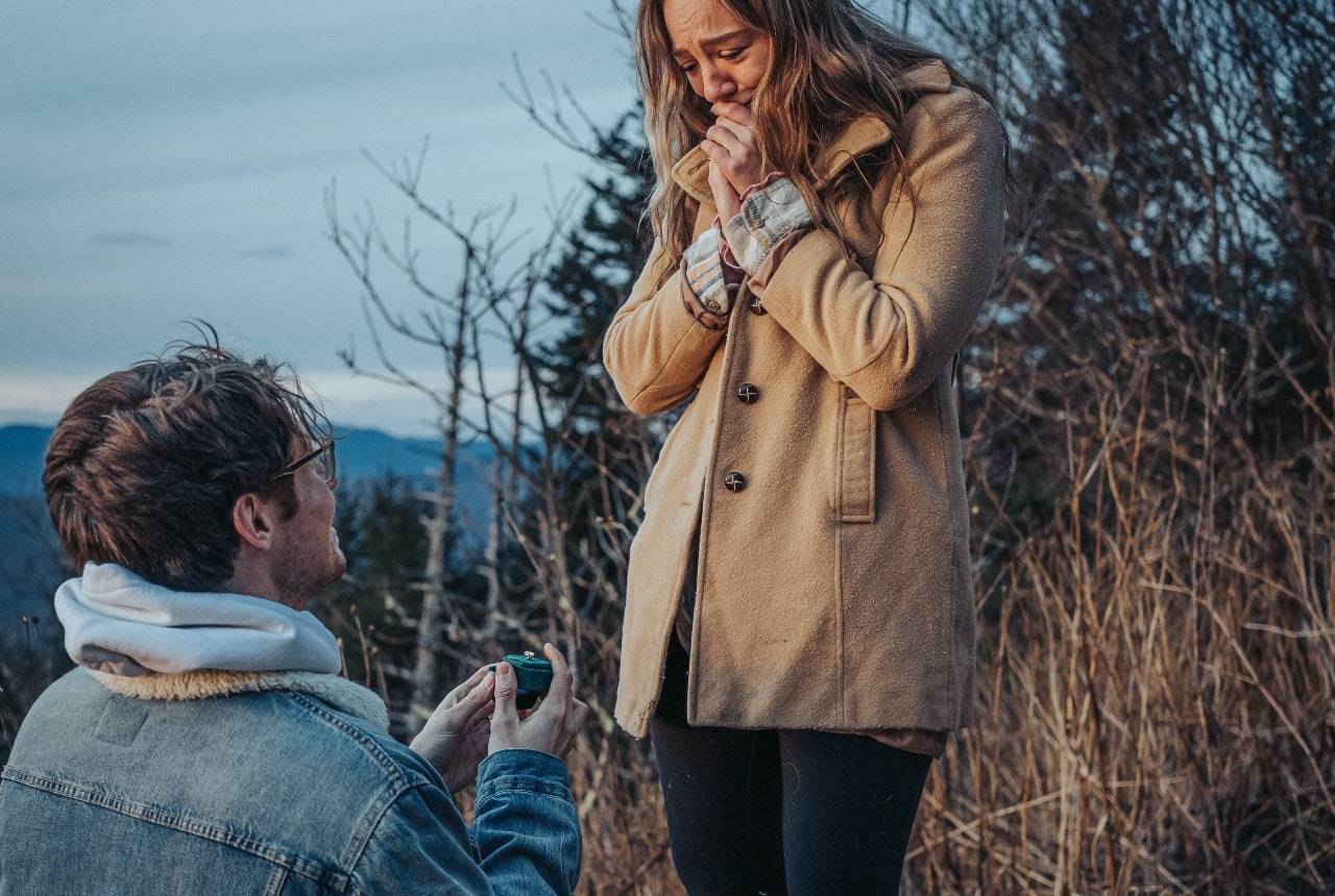 A man in a denim jacket kneels outdoors, holding a green ring box, proposing to a woman in a tan coat who looks astonished.