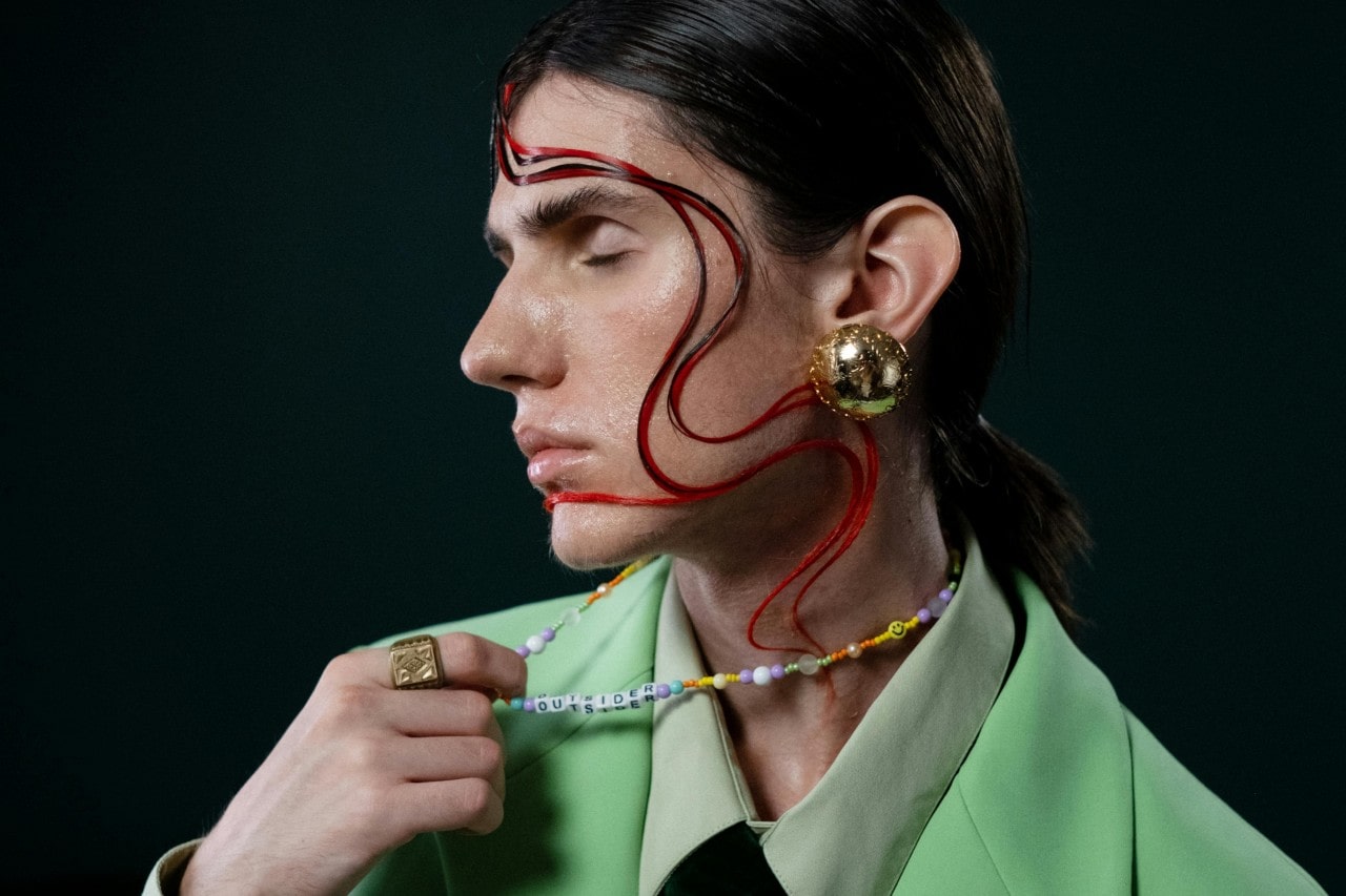 A person with a distinctive hairstyle wearing bold gold jewelry and a beaded necklace.