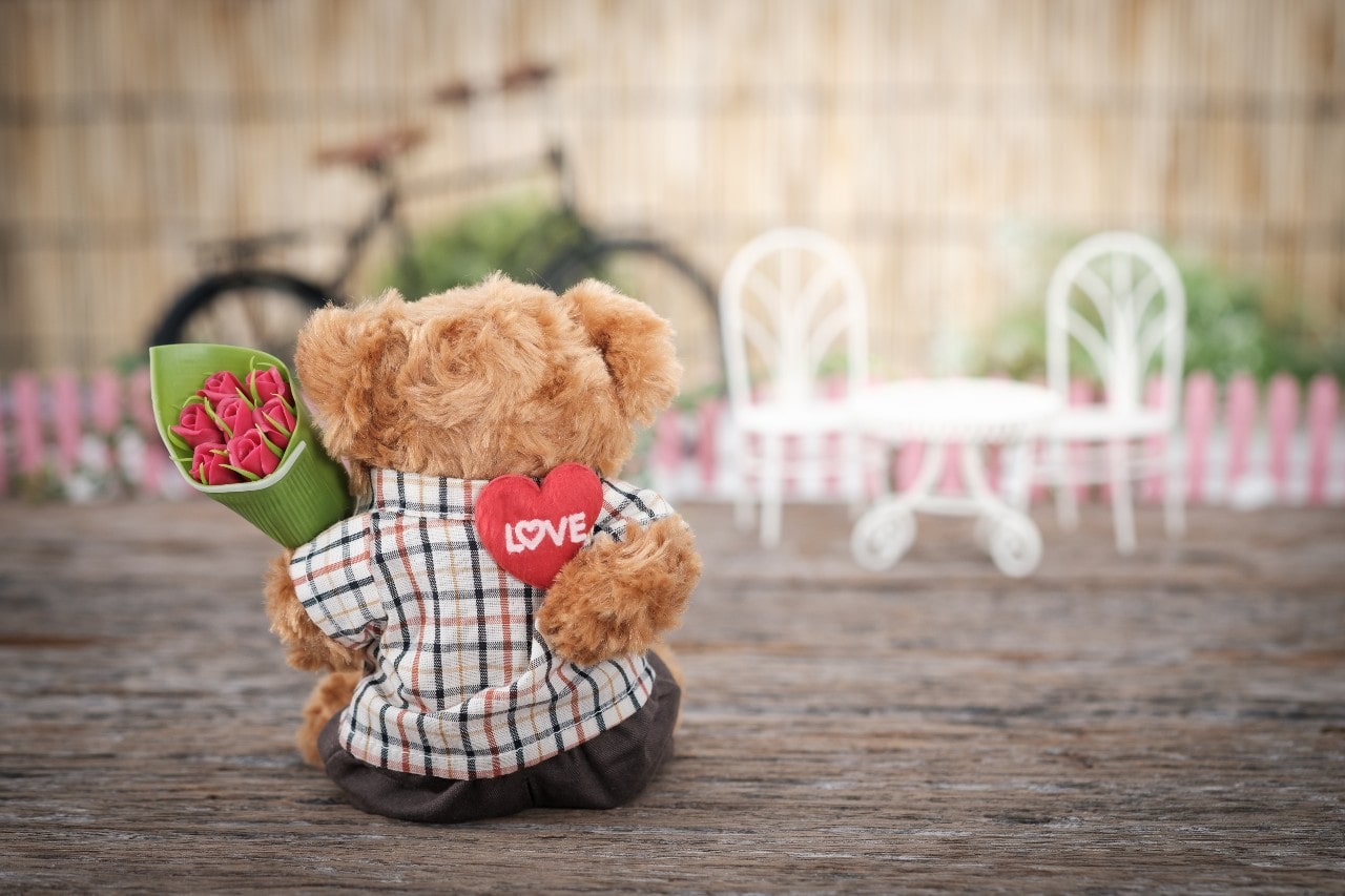 A teddy bear holds a bouquet of roses and a stuffed heart behind his back.