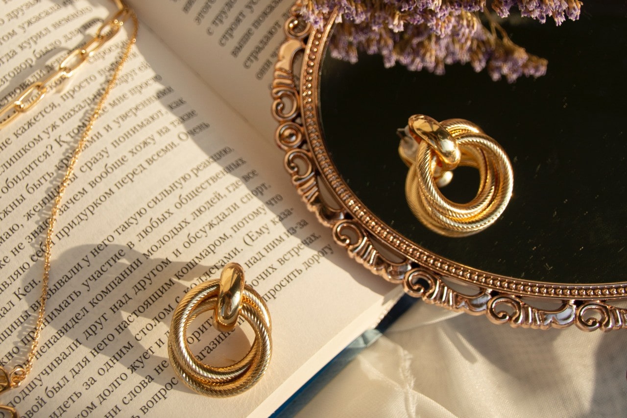 An open book with Cyrillic text lies beneath a gold chain, a pair of gold earrings, and a vintage mirror reflecting the jewelry.