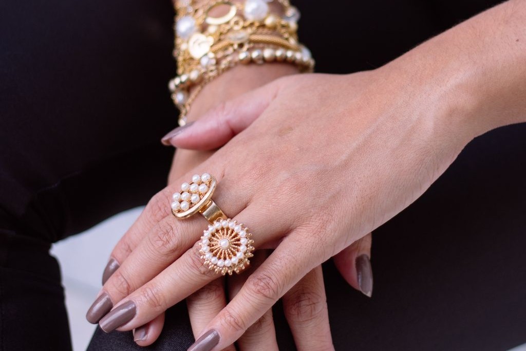 A close-up of a woman’s manicured hands resting on her lap, adorned with ornate gold rings with pearls, and layered gold and pearl bracelets on her wrists.