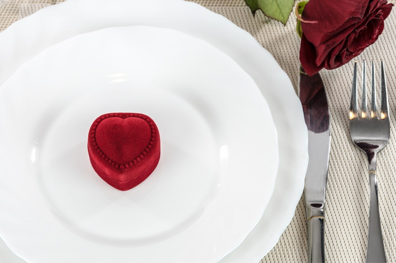A small red heartshape box of a plate at a place setting with a rose in the corner; the jewelry box would be perfect for earrings, a ring, or a pendant.
