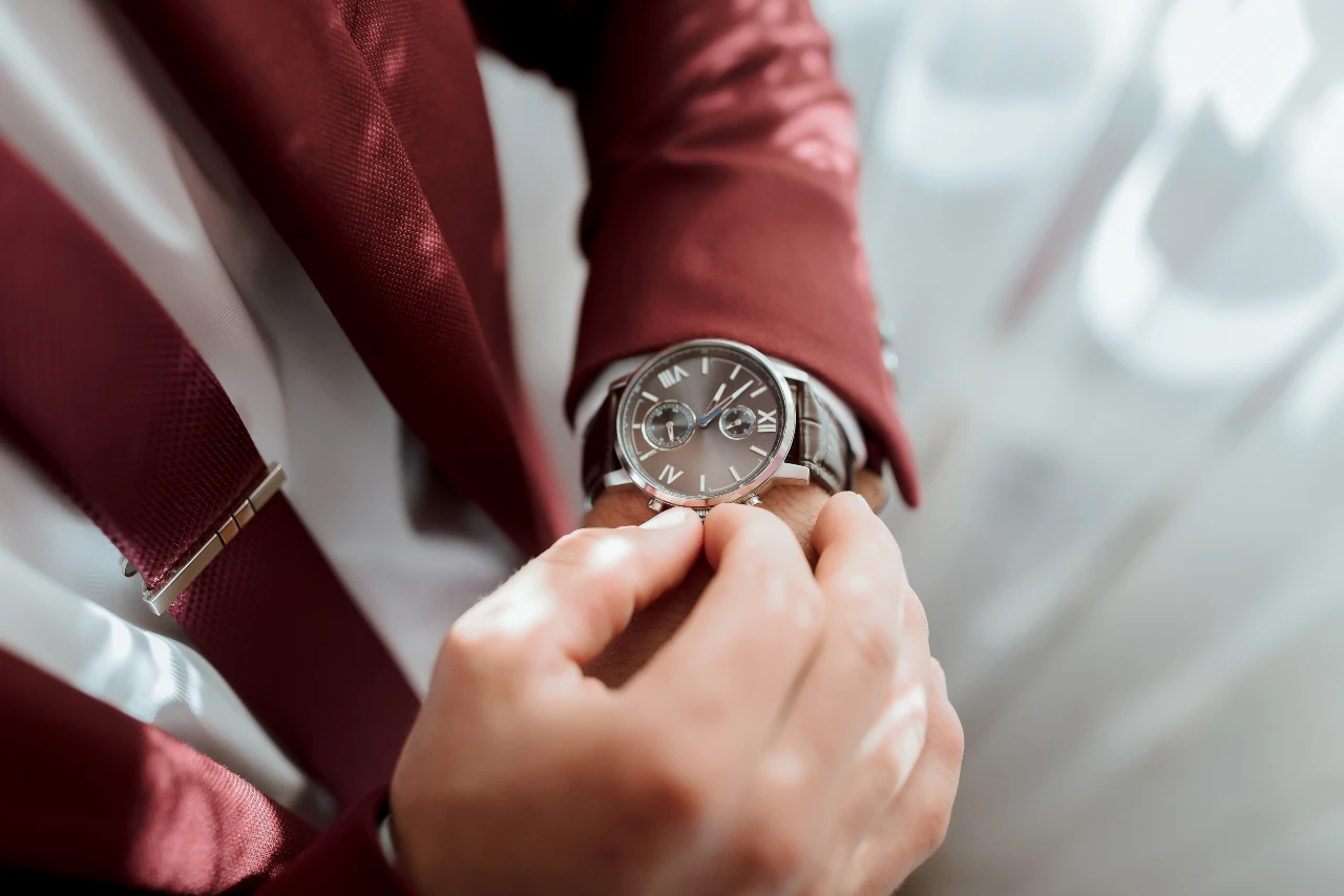 Close-up of a person in a red jacket and tie adjusting a brown leather-strapped watch on their wrist.