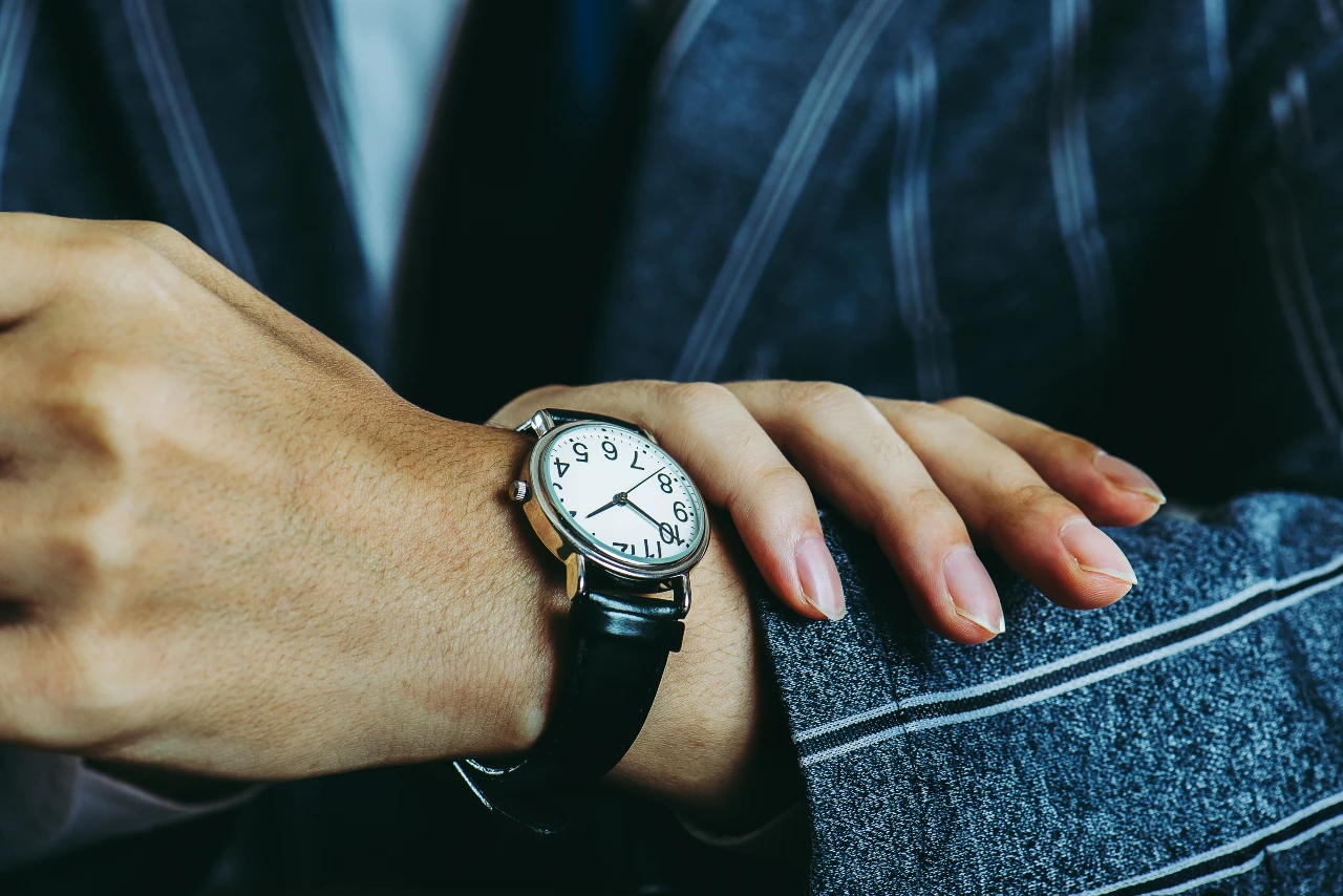 Close-up of a classic watch with black leather strap, white dial, and black numerals on a man’s wrist in a dark suit.