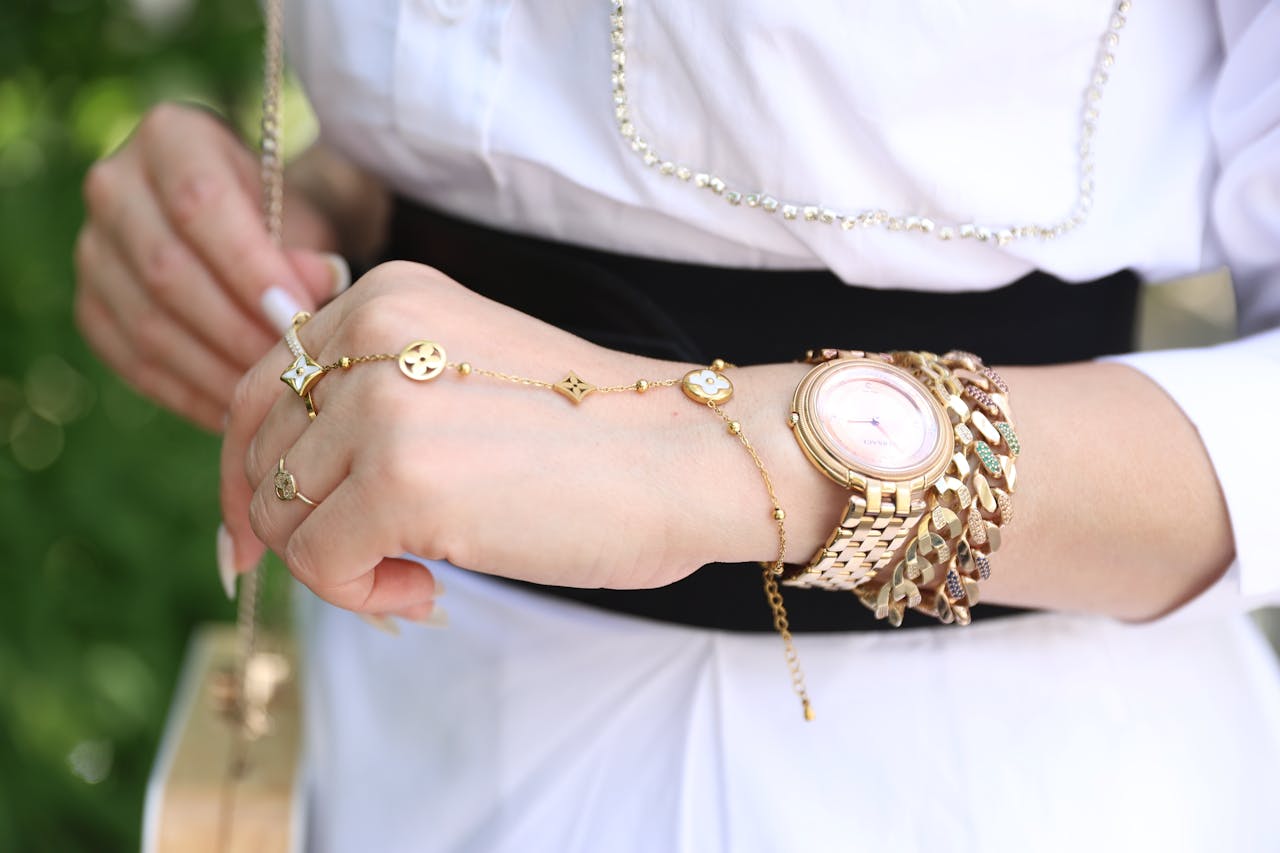 A close-up of a woman&rsquo;s forearm, her hand and wrist adorned in multiple yellow gold adornments, including a gold watch, a bold chain bracelet, and a delicate gold ring.