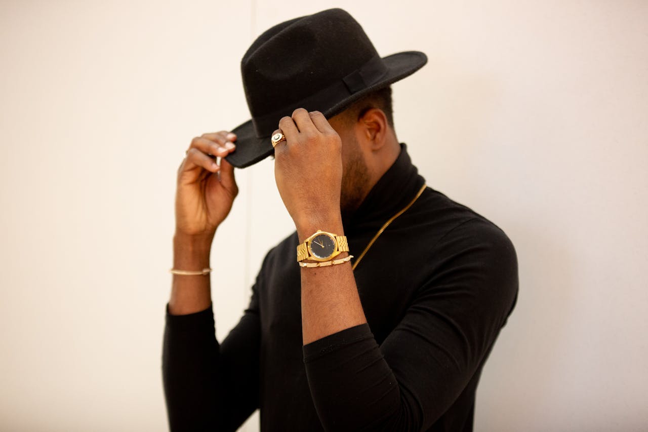 A view of a man dressed in a black turtleneck, sleek gold jewelry and a handsome gold watch obscuring his face as he adjusts the brim of his hat.